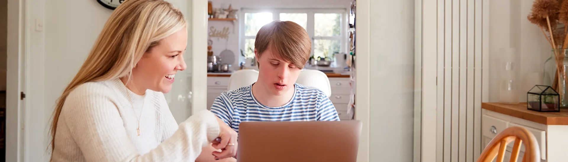A man with down syndrome is working on a laptop while a woman sitting with him helps him with tasks.