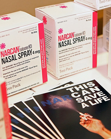 NARCAN (Naloxone HCI) Nasal Spray 4 mg" displayed on a table. The boxes are primarily white with pink and black text, and the label indicates that it is a nasal spray for reversing opioid overdoses. A pamphlet in the foreground features an image of a hand holding the spray and bold text that reads "THIS CAN SAVE A LIFE." The arrangement emphasizes the life-saving purpose of the product.