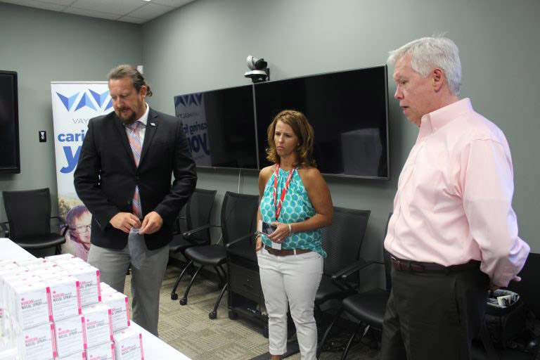 Three people, a woman and two men, standing near each other with TVs behind them looking over a table with boxes of NARCAN on it.