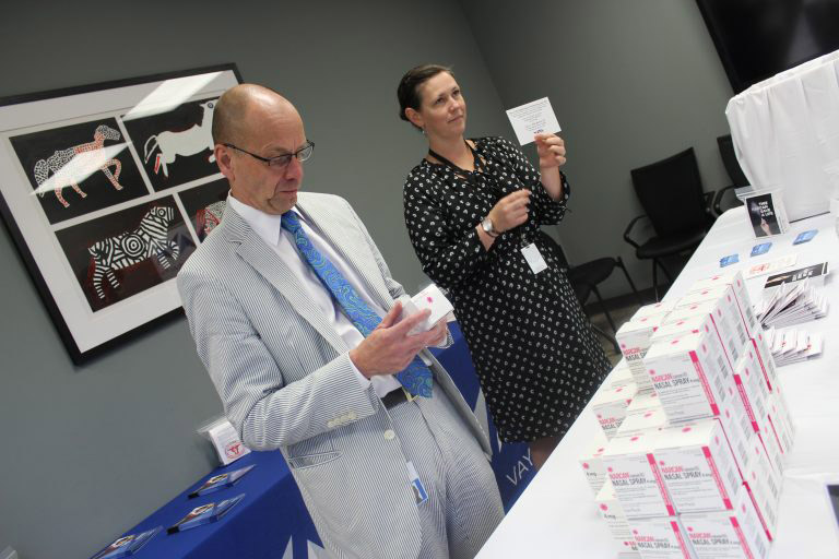 A man with a light blue suit and a darker blue tie holding a box of NARCAN standing next to a woman in a black and white dotted dress holding up a note card.