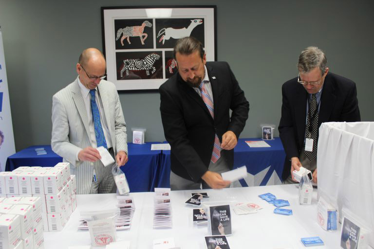 Three men in suits standing next to each other looking down at a table with boxes and flyers on it.