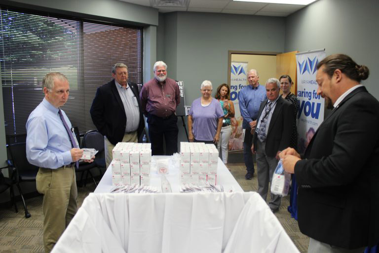A group of people standing around a table that has NARCAN on it while a man with a ponytail fills a plastic bag.