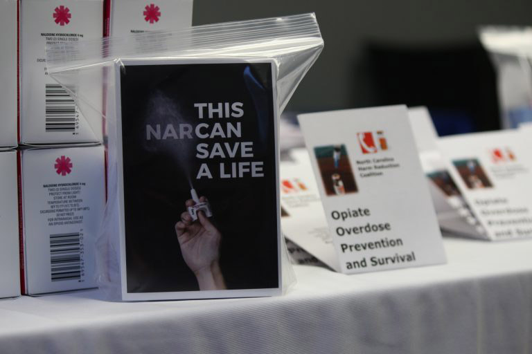 A table displaying several boxes of NARCAN with a filled plastic bag with a box in it and a flyer facing the camera saying This NARCAN save a life.