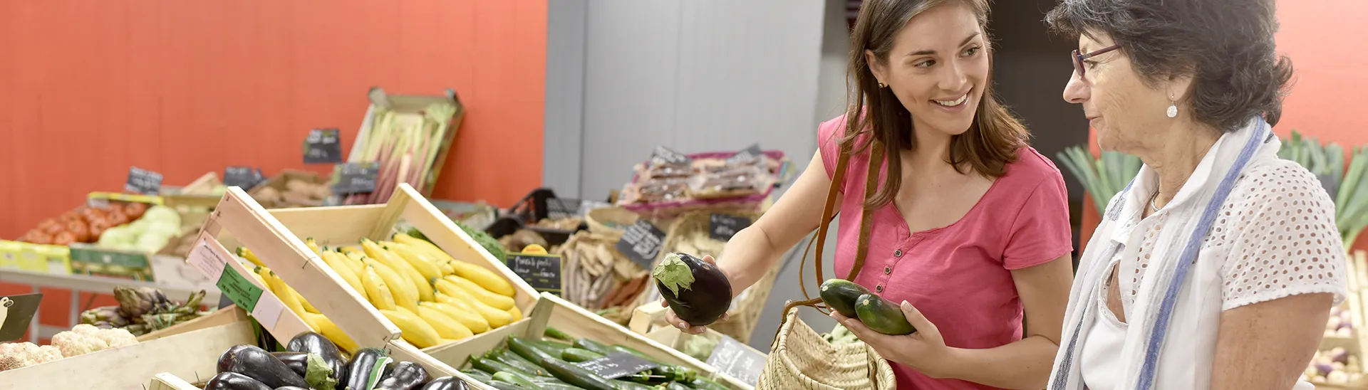 Two women talking while at a vegetable stand. One woman is holding an eggplant and two cucumbers.