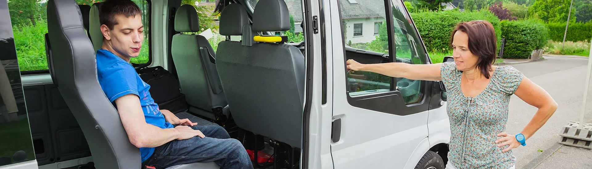 Young man, smiling while sitting in van with the doors open and woman waiting on him to exit the vehicle.