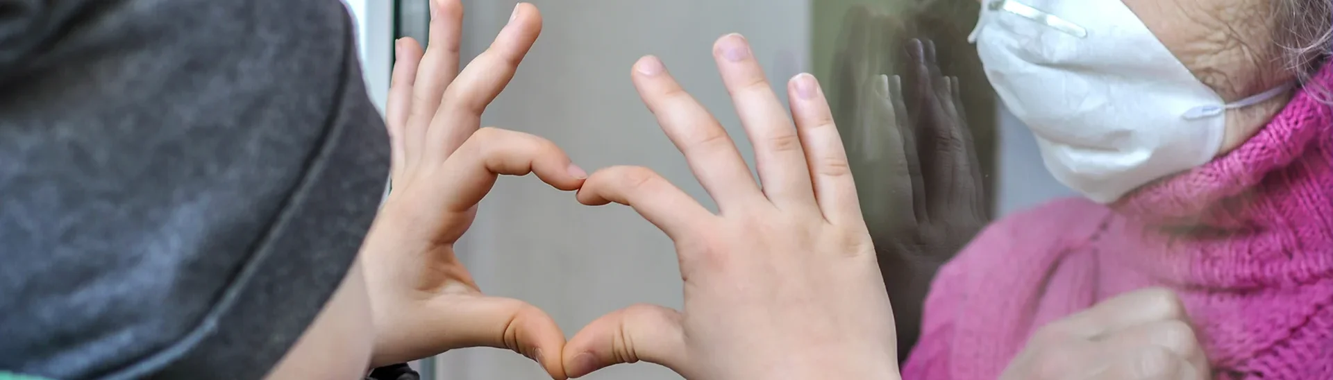 Un niño muestra su amor a su abuela durante COVID-19 levantando la mano en forma de corazón al otro lado de una ventana mientras la abuela lleva una máscara.