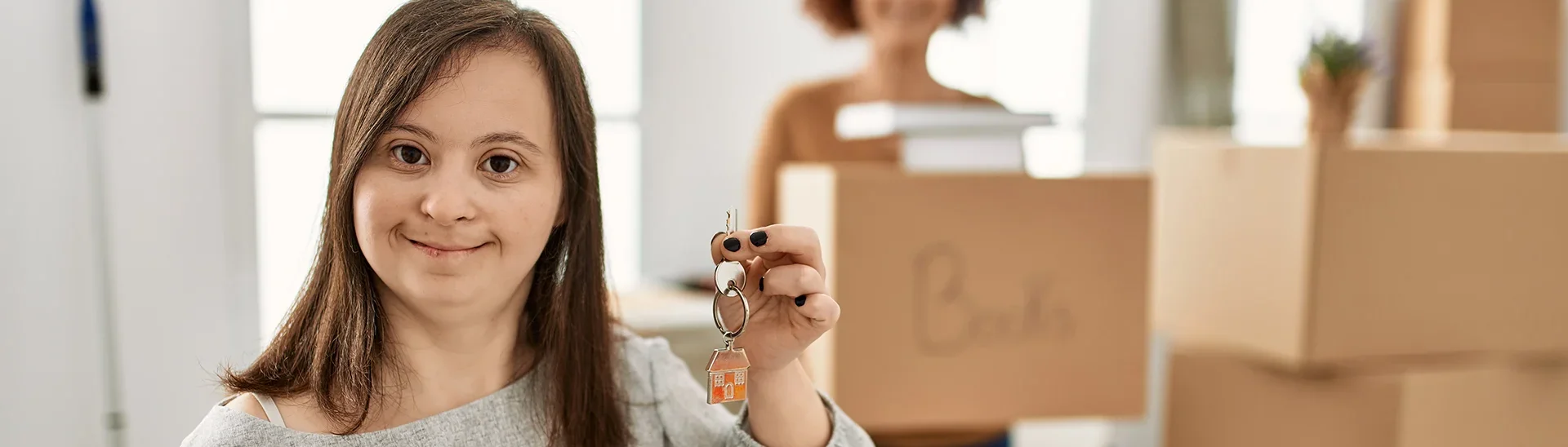 A woman with down syndrome begins moving into her own place with the help of a friend. Woman holding keys with boxes stacked behind her.