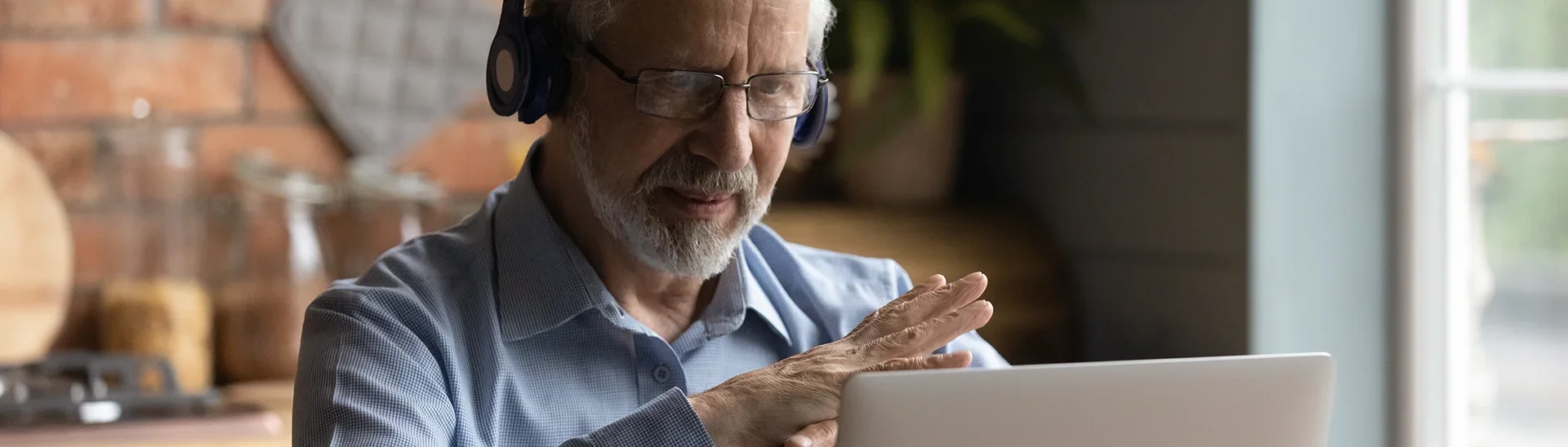 Older man with glasses and headphones on providing mentoring help virtually with his hands crossed in front of his laptop.