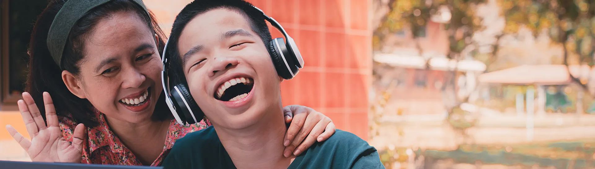 Niño con síndrome de Down en silla de ruedas con auriculares riendo, mientras una mujer sonríe con la mano alrededor de su cuello compartiendo un momento feliz.