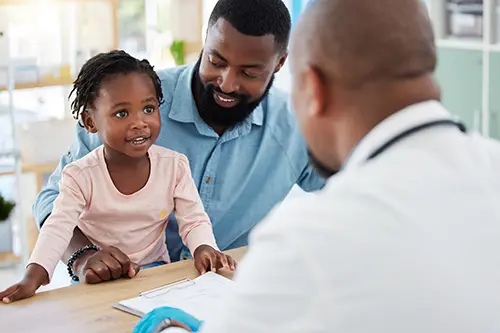 A young girl about three years old sits on the lap of her father. They are sitting across from a doctor in a white coat. 