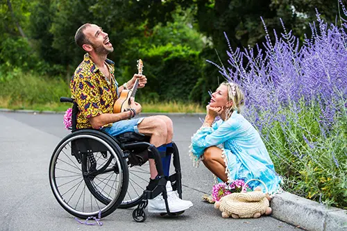 Man playing guitar and singing in a wheelchair while a woman is sitting down listening.
