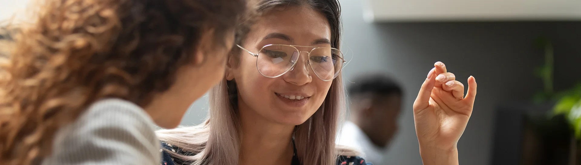 An Asian woman with glasses is sitting next to another woman smiling while they are having a conversation.