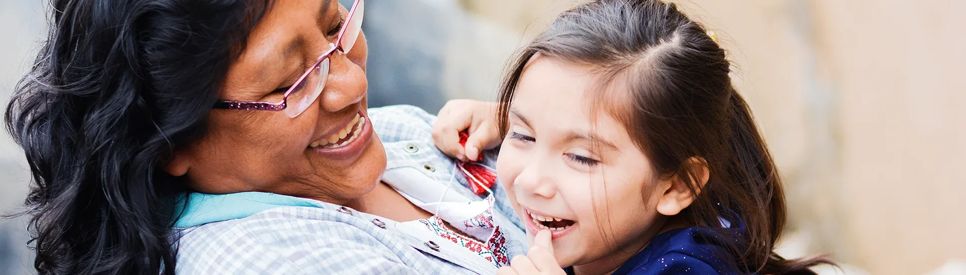 A woman with glasses and dark hair is smiling joyfully as she holds a young girl in her arms. The girl, wearing a navy blue outfit, is laughing with her eyes squinted and hand near her mouth. The background is softly blurred, highlighting their happiness and the close bond they share.