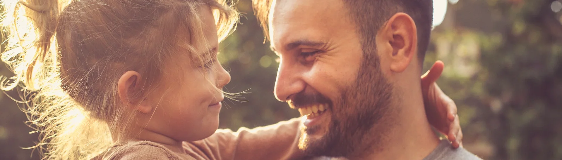 A father with a beard and short hair is holding his young daughter in his arms. They are smiling warmly at each other, sharing a loving gaze. The little girl, with her hair in a ponytail and wearing a brown sweater, wraps her arm around his neck. The background features soft sunlight and blurred trees, creating a golden, heartwarming atmosphere.