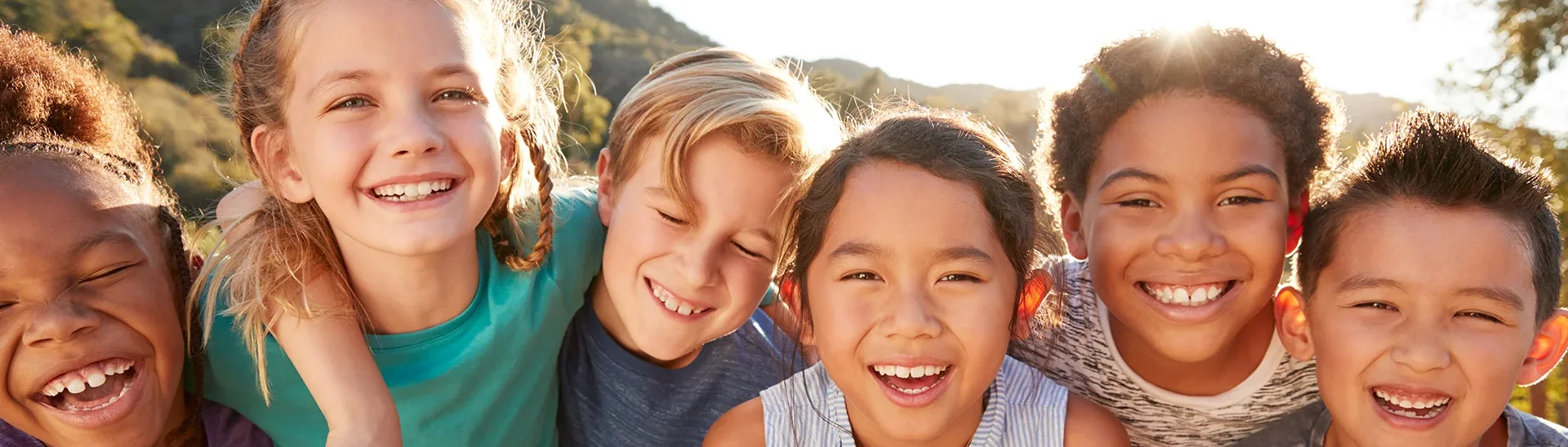 Un grupo de seis niños diversos está de pie, muy juntos, al aire libre, sonriendo y riendo mientras se inclinan hacia la cámara. Van vestidos con ropa informal y la luz del sol proyecta un cálido resplandor sobre sus rostros. El fondo está formado por colinas y árboles, lo que sugiere un entorno natural al aire libre. Sus expresiones transmiten felicidad, amistad y alegría.