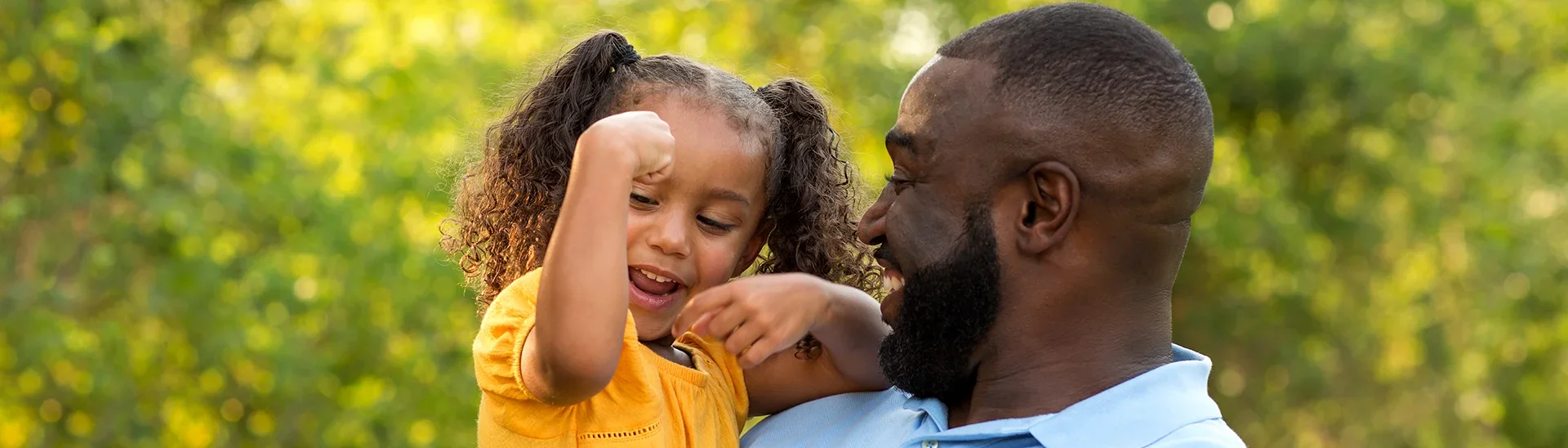 A father with a beard and short hair is holding his young daughter outdoors in a sunny, green setting. The little girl, wearing a yellow shirt and with curly pigtails, is playfully flexing her arm while smiling. The father looks at her with a joyful and loving expression, capturing a moment of bonding and happiness.