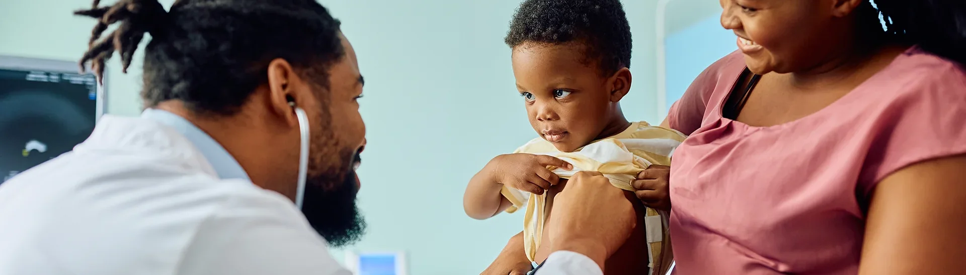 A young child is sitting on their mother’s lap in a medical examination room, while a doctor with a stethoscope listens to the child’s chest. The child, wearing a yellow shirt, looks slightly curious and cautious, while the mother smiles warmly. The doctor, with a friendly demeanor, focuses on the examination. The setting is clean and brightly lit, creating a caring and reassuring atmosphere.