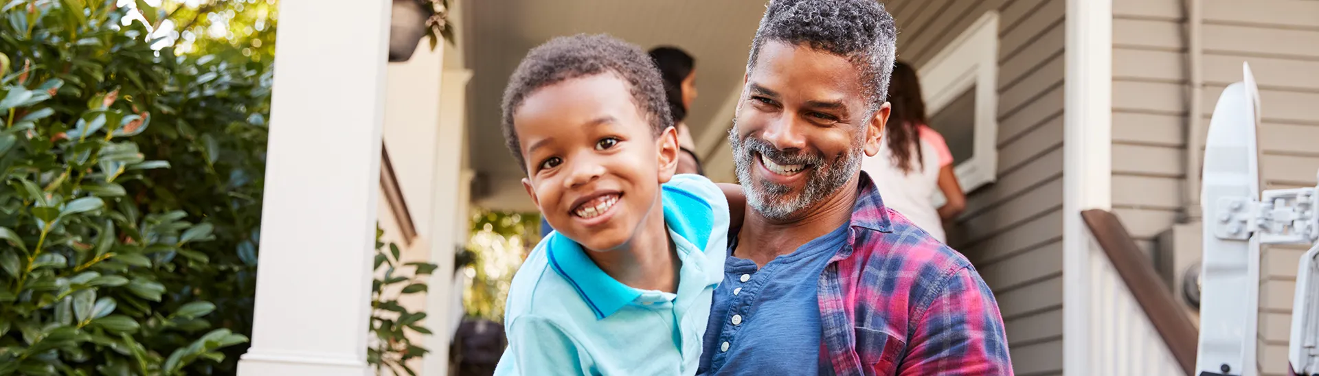 Un hombre sonriente, con pelo rubio y barba, sostiene a un niño en brazos. El niño, que lleva una camisa azul brillante, sonríe alegremente y mira a la cámara. Están de pie en el porche de una casa con el exterior gris, columnas blancas y vegetación de fondo, lo que confiere a la escena un ambiente familiar cálido y alegre.