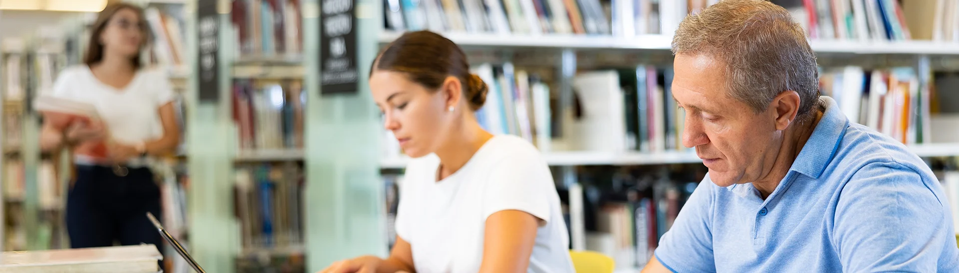 An older man in a light blue polo shirt is sitting at a table in a library, writing on a notebook while using a laptop. Next to him, a younger woman in a white shirt is also focused on her work, typing on her laptop. In the background, bookshelves filled with books and another woman holding a book can be seen, creating a studious and quiet atmosphere.