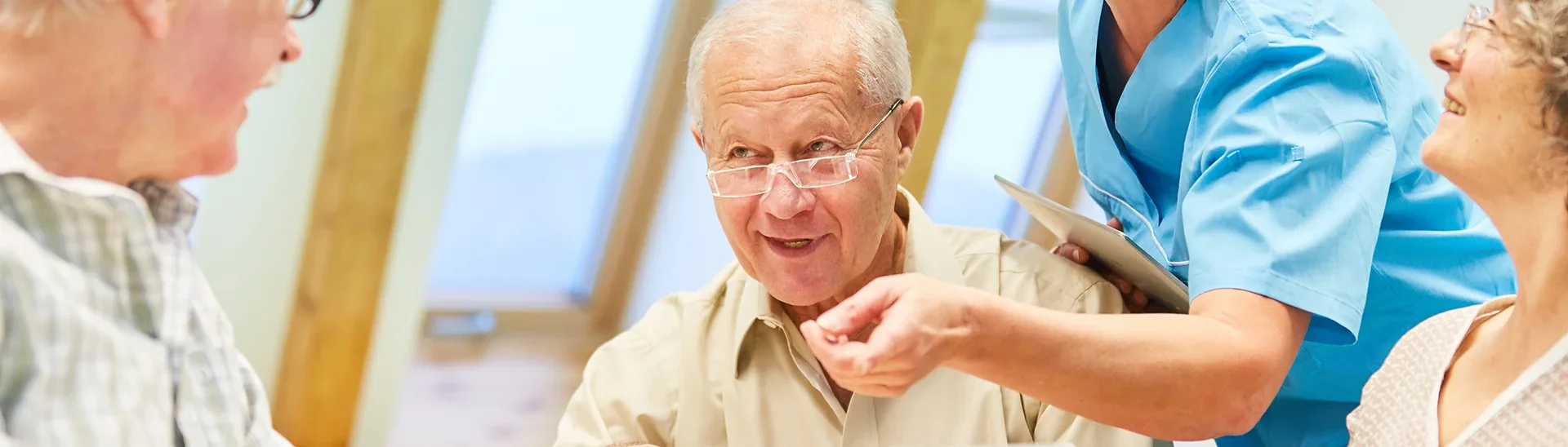 Un profesional sanitario sonriente, vestido con una bata azul, atiende a un grupo de ancianos sentados a una mesa. El grupo, formado por tres ancianos, sostiene tabletas e interactúa entre sí. El entorno es una sala moderna y luminosa con grandes ventanales y luz natural, lo que crea un ambiente cálido y positivo centrado en el aprendizaje o la conexión.