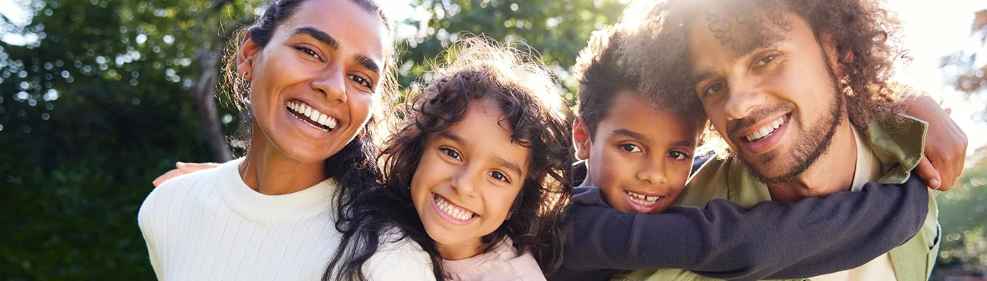 Una sonriente familia de cuatro personas posa al aire libre en un entorno soleado y verde. Los padres, una mujer con jersey crema y un hombre con chaqueta verde, están muy juntos con sus dos hijos, que les abrazan juguetonamente por detrás. Los niños, un chico y una chica de pelo rizado, sonríen alegremente. La luz del sol se cuela entre los árboles, dando a la escena un ambiente cálido y alegre.
