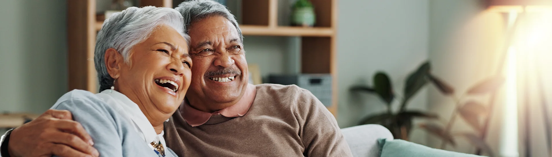 An elderly couple with gray hair is sitting together on a couch, smiling and laughing. The man has his arm wrapped around the woman, and they both appear happy and relaxed. The background features a cozy living room with a bookshelf, plants, and soft lighting.