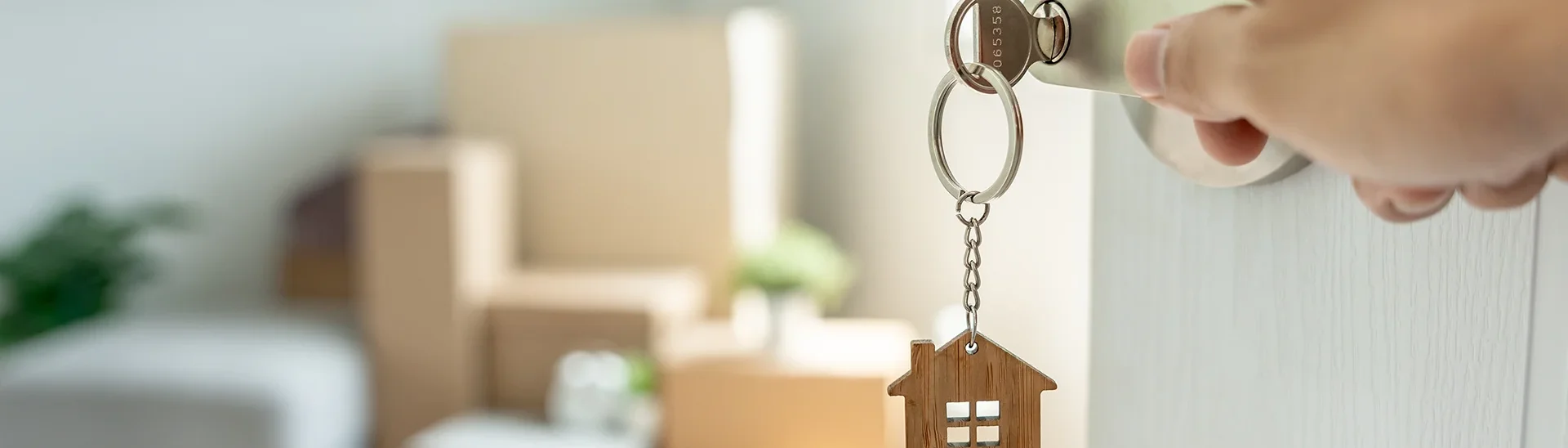 Key handing from the lock of a home's door opening to a blurry image of boxes sitting in a room.