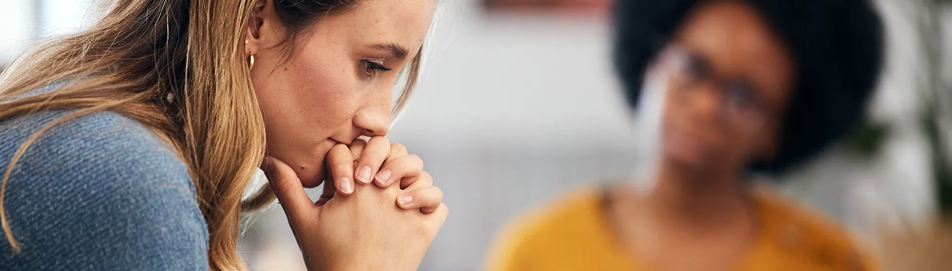 A woman looking down with her hands covering her both, in deep thought while a woman with glasses looking on caringly in the background.