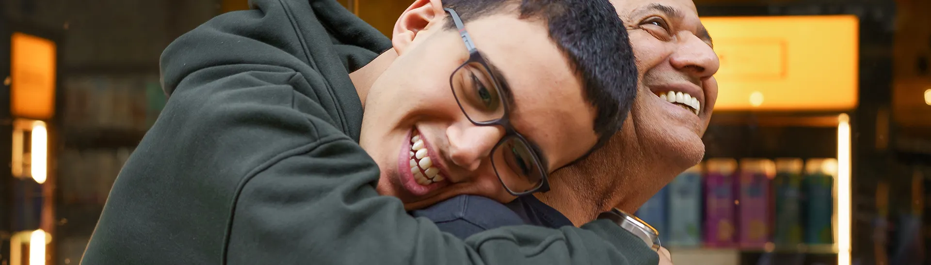 Un hijo con gafas abraza a su padre por detrás mientras ambos sonríen juntos.