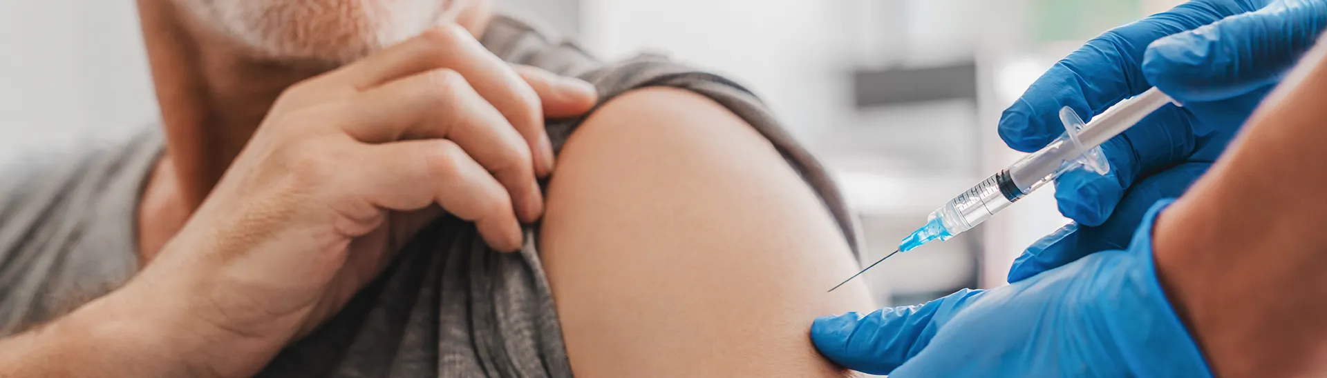 Un hombre recibe una inyección de vacuna en el hombro mientras sujeta la manga mientras el médico con guantes azules le introduce la aguja en el brazo.