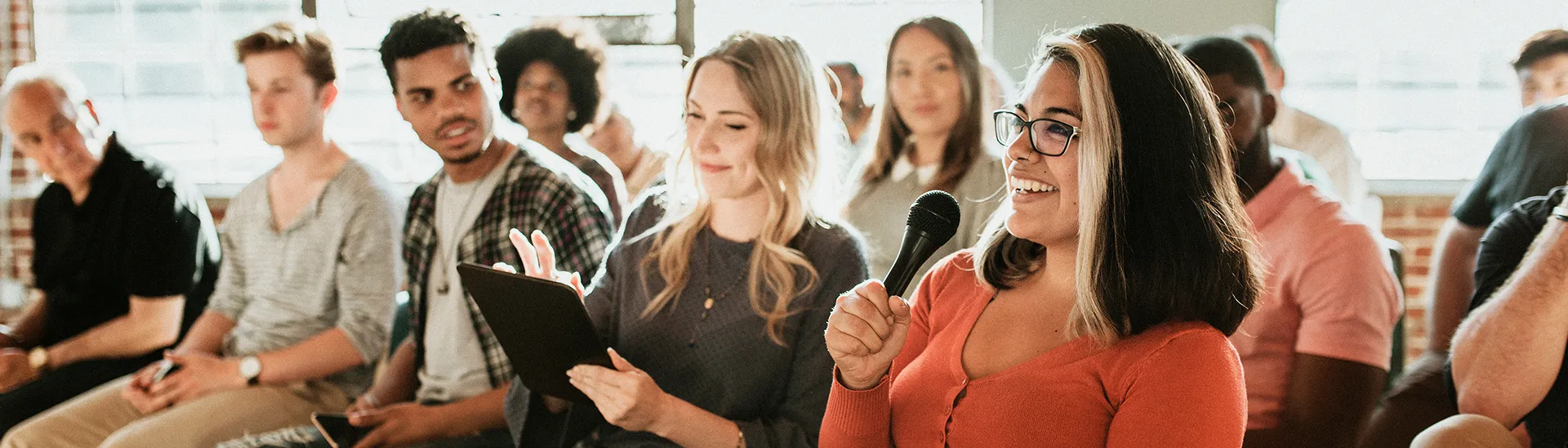 A mixed group of individuals attending a community meeting and actively participating as one woman holds a microphone smiling during the conversation.