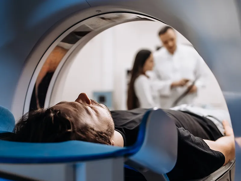 Man-In-CT-Scan-Machine_2519637569 Un hombre yace dentro de una máquina de tomografía computarizada mientras dos médicos hablan y revisan su historial.