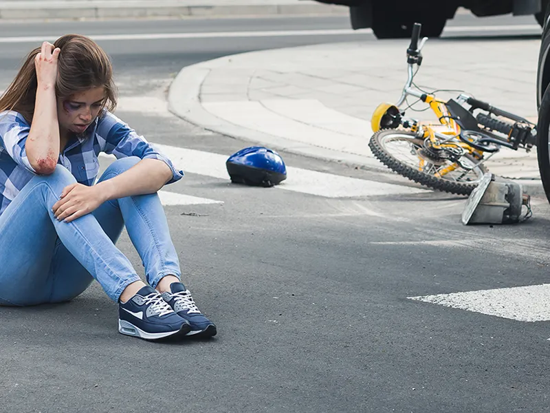 Woman-Bicycle-Accident-Head_581251246 Una mujer sentada, sujetándose la cabeza, tras ser atropellada por un coche mientras montaba en bicicleta.