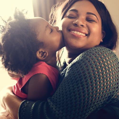 A little girl kisses her mother's cheek and the mother is smiling brightly for it.