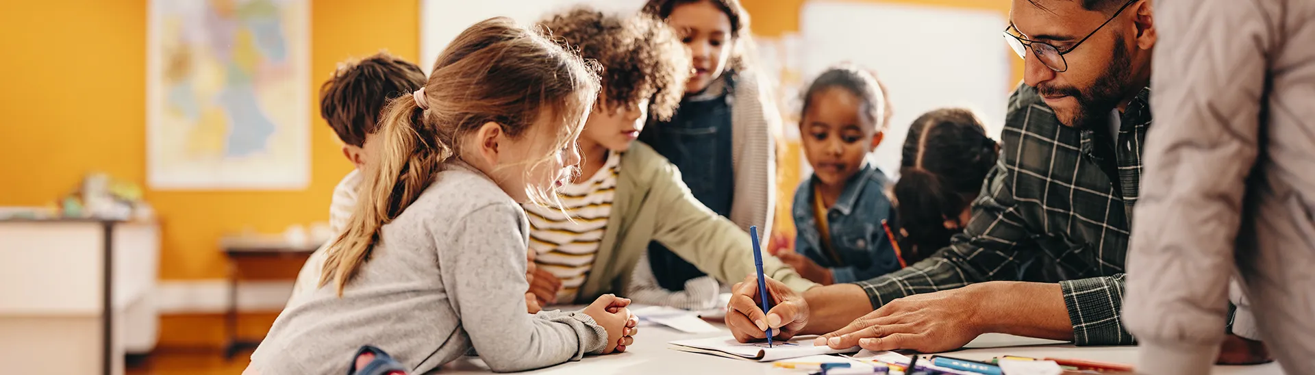 A teacher is writing on a workbook while younger students are gathered around him watching what he does.