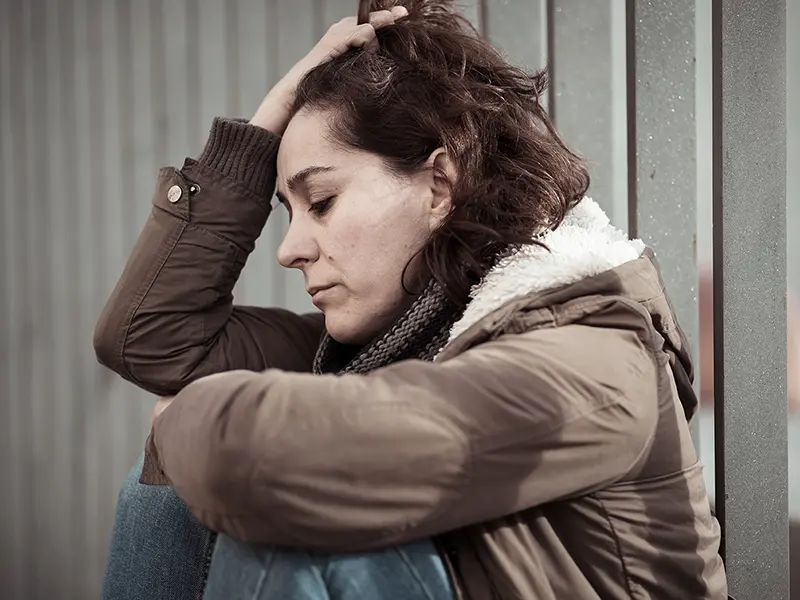 Una mujer con un abrigo de invierno sentado contra una barandilla con la mano en el pelo con aspecto angustiado.